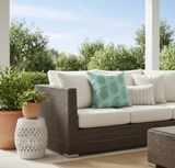Outdoor wicker sofa with neutral cushions, accented by a green-blue pillow with paisley patterns and a striped cream pillow, set on a covered patio with potted plants.