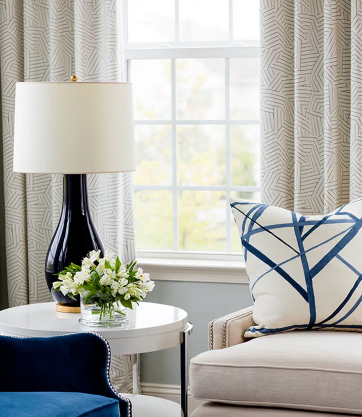Cream-colored armchair with a white pillow featuring bold navy blue abstract lines, next to a round white side table holding a navy blue lamp and small floral arrangement.