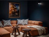 Tan sectional sofa with gray and white geometric patterned throw pillows and a white throw blanket in a dark blue-walled room with a colorful abstract painting and round wooden stools.