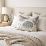Beige bed with a floral pillow featuring blue and green Kalamkari designs, flanked by plain cream pillows and a textured beige throw blanket at the foot.