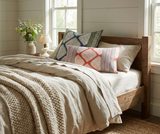 Bed with light beige linen bedding, chunky knit blanket, and three patterned pillows including a rectangular one with red geometric design and two square ones in blue and green tones.