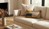 Rectangular pillow cover with color blocks in tobacco brown, teal, and beige on a cream sofa next to a window with sheer curtains.