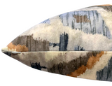 Close-up of a beige velvet pillow with abstract brush stroke patterns in blue, brown, and gray, featuring a discreet zipper along the edge.