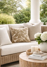 Outdoor woven rattan sofa with beige cushion and assorted neutral-toned pillows, including a leaf-patterned pillow, beside a round wooden table with candles and white flowers.