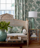 Vintage-style wooden settee with beige cushion, sage floral pillow and bolsters, set against matching floral wallpaper and curtains, beside a green glass lamp on a side table.