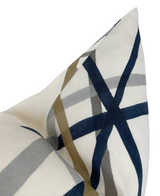 Close-up of a pillow cover with intersecting navy blue, taupe, and gray brushstroke lines on a sand-colored fabric background, highlighting a pointed corner.