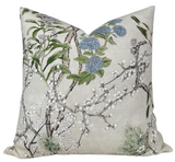 Square beige fabric pillow with a botanical print featuring white blossoms, blue flowers, and green leaves on thin brown branches.