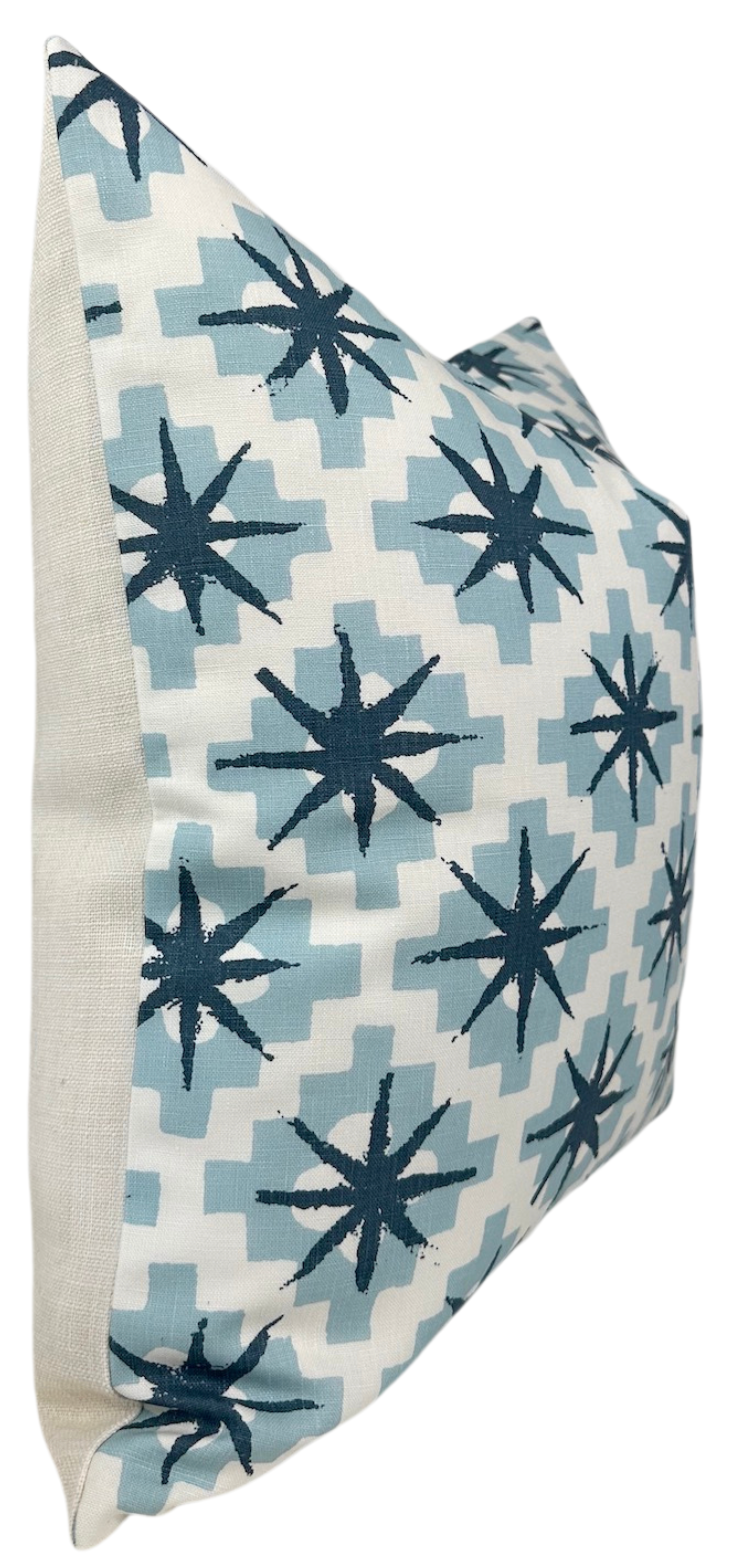 Square pillow with a pattern of dark blue starbursts over light blue geometric shapes on a white background, displayed at an angle showing a cream side panel.