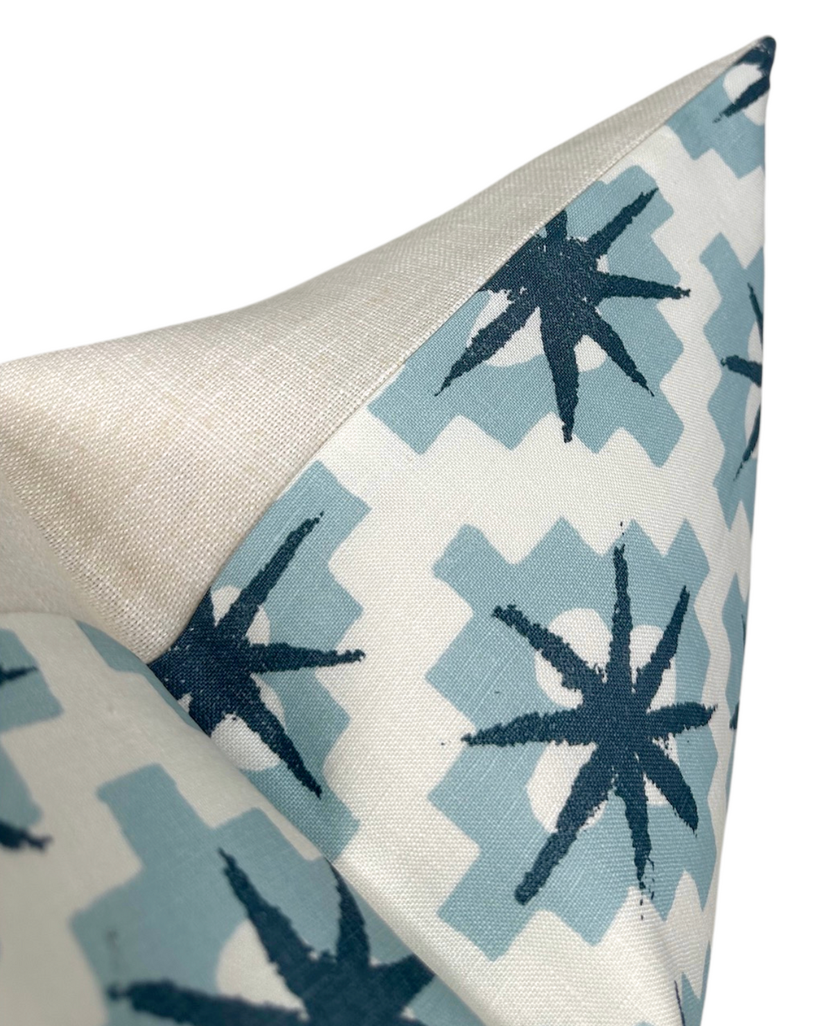 Corner of a pillow cover featuring dark blue starburst shapes on muted blue geometric blocks with a beige woven fabric on the reverse side.
