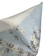 Corner detail of a pillow with light blue fabric featuring white and yellow weeping blossom flowers and gray leaves, backed by a textured beige fabric.