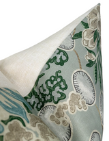 Close-up of a sage-colored pillow cover showing detailed floral and leaf patterns in green, beige, and gray tones on textured fabric beneath a folded natural linen corner.
