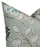 Close-up of a sage green pillow cover with embroidered floral and leaf patterns in shades of white, blue, black, and beige on a textured fabric.
