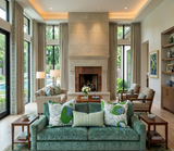 Spacious living room with a green velvet sofa adorned with floral and striped cushions, centered in front of a large stone fireplace framed by tall windows and beige curtains.