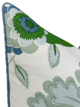 Close-up of a decorative pillow corner featuring green and blue floral embroidery on white fabric with blue piping along the edge.
