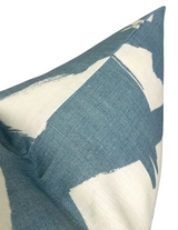 Close-up of a linen pillow cover featuring bold, irregular sky blue brushstroke patterns on a natural off-white background with visible fabric texture.