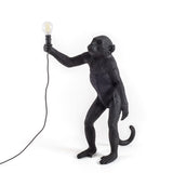 Black standing monkey sculpture lamp holding an exposed filament bulb in its raised right hand, with a visible black electrical cord hanging downward.