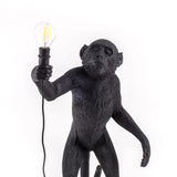 Black sculpted monkey lamp standing upright, holding a light bulb in its raised right hand against a white background.