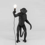 Black standing monkey sculpture holding a lit lightbulb in its right hand with a cord hanging loosely, shown against a plain white background.
