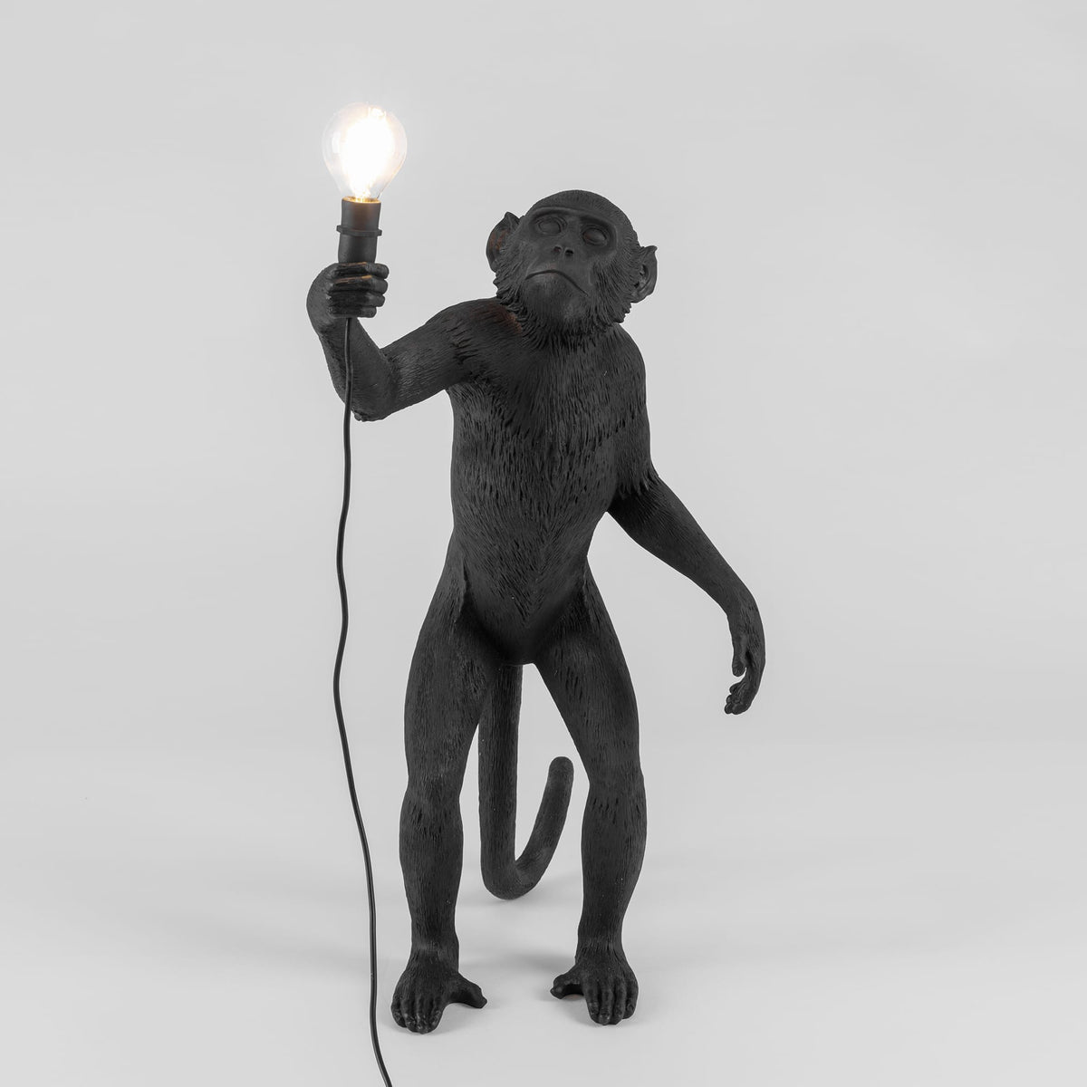 Black standing monkey sculpture holding a lit lightbulb in its right hand with a cord hanging loosely, shown against a plain white background.