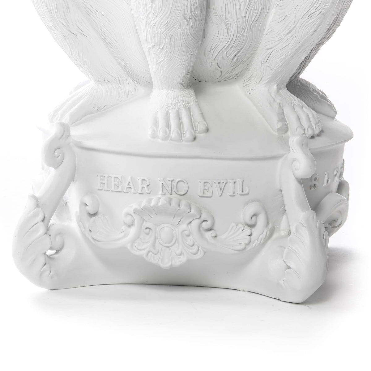White sculpted pedestal with detailed ornate scrollwork and the inscription 'HEAR NO EVIL' beneath seated figure's feet with visible toes and nails.