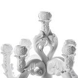 White sculpted chandelier-like object with six ornate candle holders featuring leaf and scroll motifs, arranged symmetrically around a central stem, set against white background.