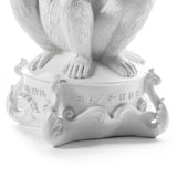White sculpture base with detailed curled motifs and embossed text including 'NO EVIL' and 'S L T * M X', supporting a figure with textured feet and legs.