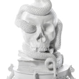 White sculpture of a large skull with a textured snake coiled around and over it, resting on a stack of books with visible pages and ornate base details.