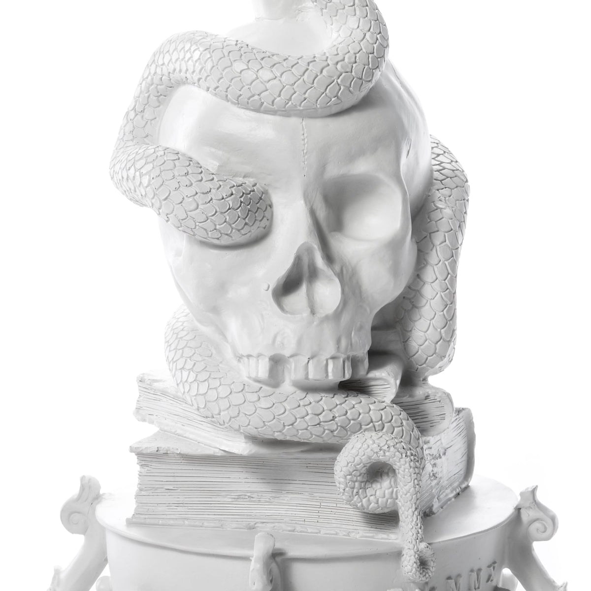 White sculpture of a large skull with a textured snake coiled around and over it, resting on a stack of books with visible pages and ornate base details.