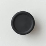 Matte black circular coat hook with a slightly recessed center, mounted on a white textured wall, showing a sleek minimalist design.