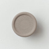 Circular matte beige Sequence coat hook with a slightly recessed center and subtle speckle texture, mounted on a white wall, shown from the front.