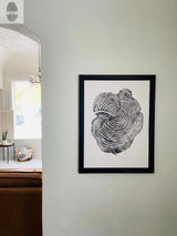 Black and white print of a detailed cross-section of a tree trunk in a black frame hanging on a textured white wall near an archway leading to a bright room.