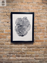 Black and white print of a tree cross-section showing detailed growth rings, framed in black and hung on a textured light brown brick wall.