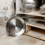Single stainless steel frying pan with a polished interior leaning against a rustic textured wall beside a wood shelf holding stacked pans.
