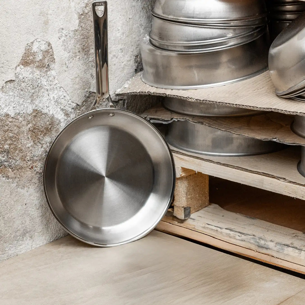 Single stainless steel frying pan with a polished interior leaning against a rustic textured wall beside a wood shelf holding stacked pans.