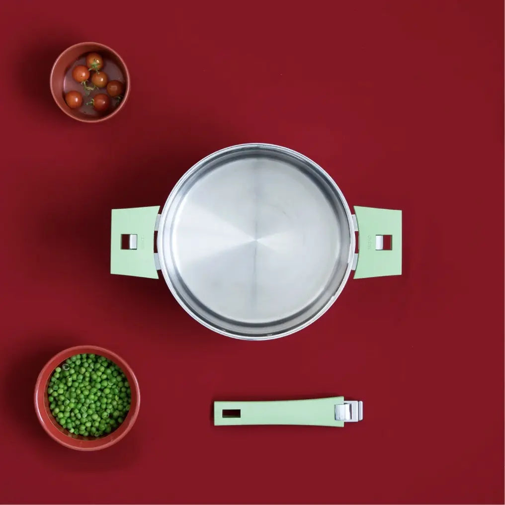 Stainless steel pan with square mint green handles on a red background, accompanied by a matching handle piece and bowls of cherry tomatoes and green peas