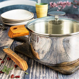 Stainless steel Cristel saucepan with lid features warm-toned wood handles set on rustic wooden surface beside stacked white plates and a green cup.