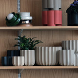 Set of four round wrap planters in two colors—beige and black—with vertical ridges, displayed on wood shelves with a small green plant in one beige planter.