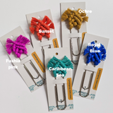 Five metal bookmarks topped with knotted macrame tassels in fuchsia pink, sunset orange, ochre yellow, Caribbean sea turquoise, and royal blue, each on a white card backing.
