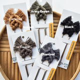 Six macrame bookmarks with knotted patterns and fringed ends in caramel, sunset, army green, black, taupe, and charcoal, displayed on white cards atop a round wooden tray.