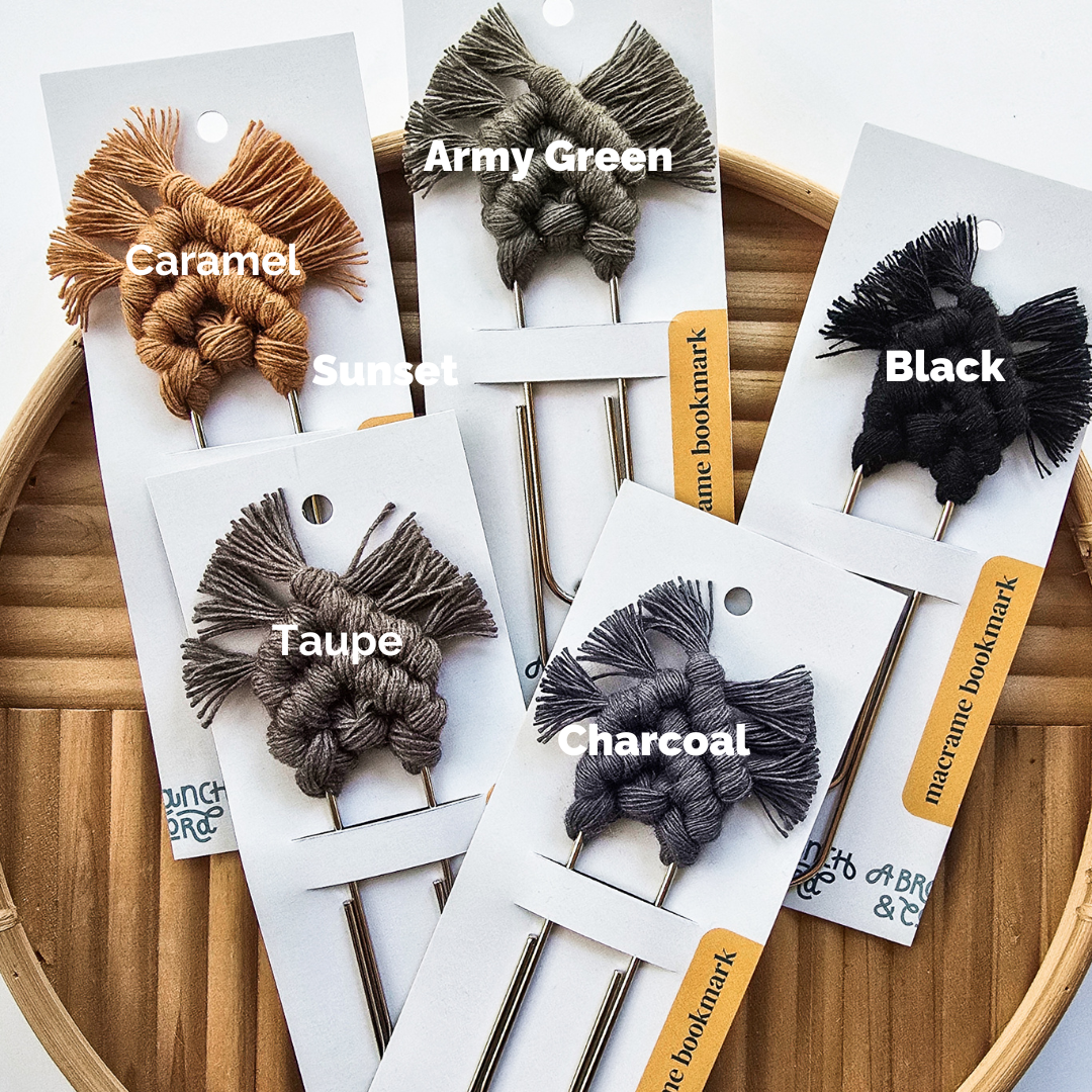 Six macrame bookmarks with knotted patterns and fringed ends in caramel, sunset, army green, black, taupe, and charcoal, displayed on white cards atop a round wooden tray.