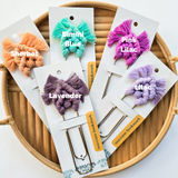 Five macrame bookmarks with fringed, fan-shaped tops in sherbet orange, bimini blue, pink lilac, lavender, and lilac shades arranged on a round wooden tray.