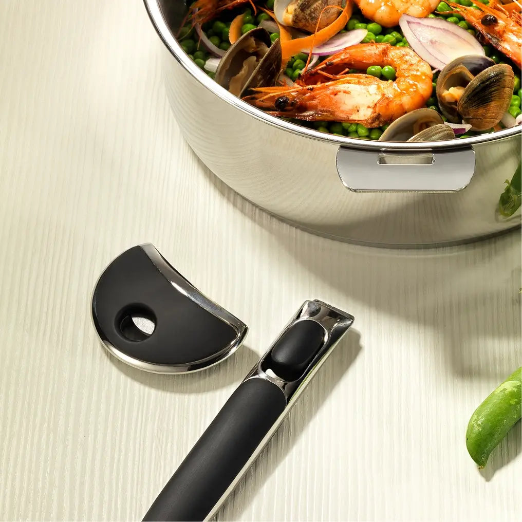Close-up of a stainless steel pot filled with shrimp, clams, green peas, and red onion slices, alongside a detachable black and silver handle on a light wood surface.