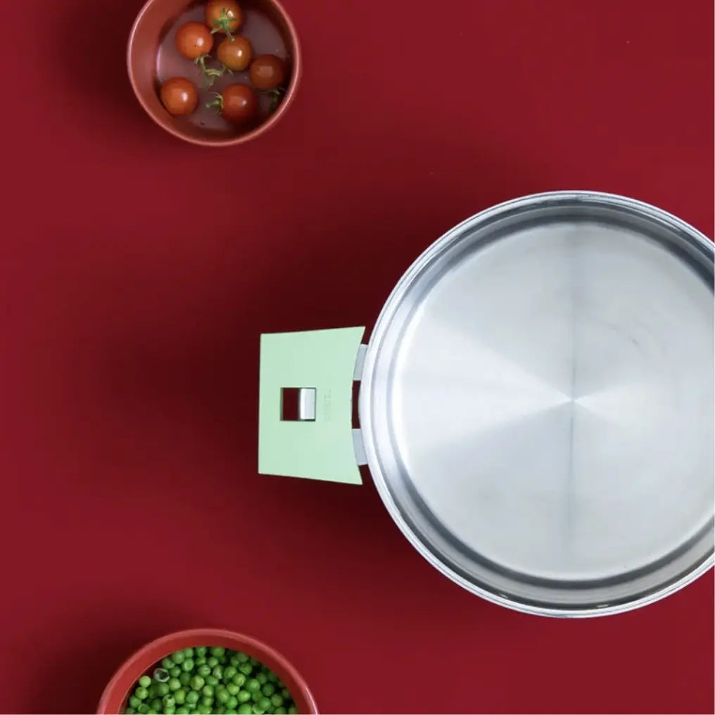 Top-down view of a stainless steel pot with a pale green side handle and round red bowls containing cherry tomatoes and green peas on a red surface.