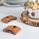 Two curved wooden handle attachments with metal inserts resting on a white surface near a stainless steel pot on a bark-edged wooden trivet.