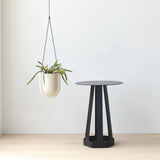 Black metal side table with a round top and six-legged hexagonal base on light wood floor against a white wall, next to a hanging white ceramic planter with green trailing plants.