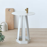 Small white metal side table with hexagonal base and flat round top, featuring a brass valve handle on center, placed on light wood surface with blurred decor background.