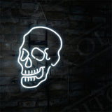 White neon light shaped like a stylized skull hanging on a dark brick wall with a metal chain, illuminating the contours and teeth details.