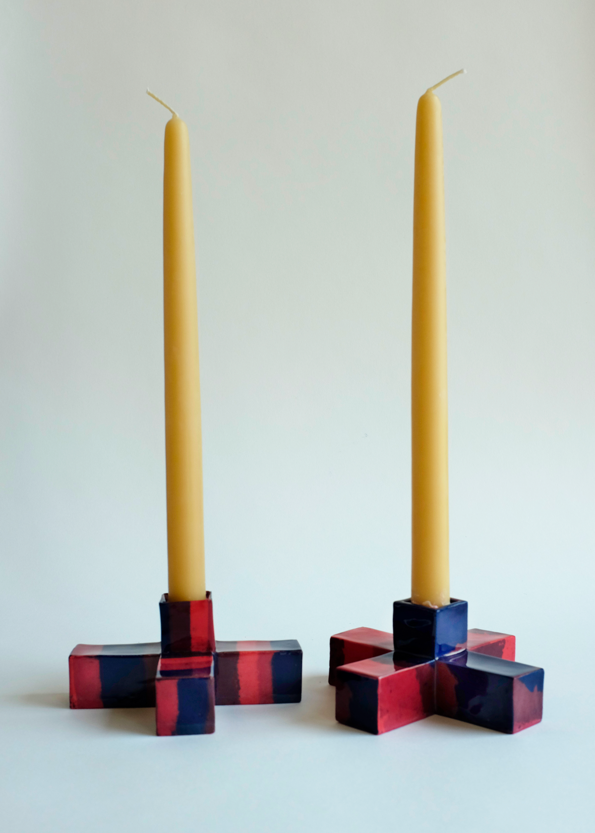 Two square-shaped candle holders with interlocking red and blue resin blocks, each holding a tall, slender yellow taper candle on a white background.