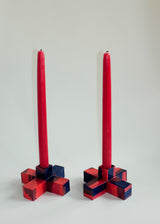 Two small squared candle holders in red and dark blue geometric patterns, each holding a tall red tapered candle against a plain light background.