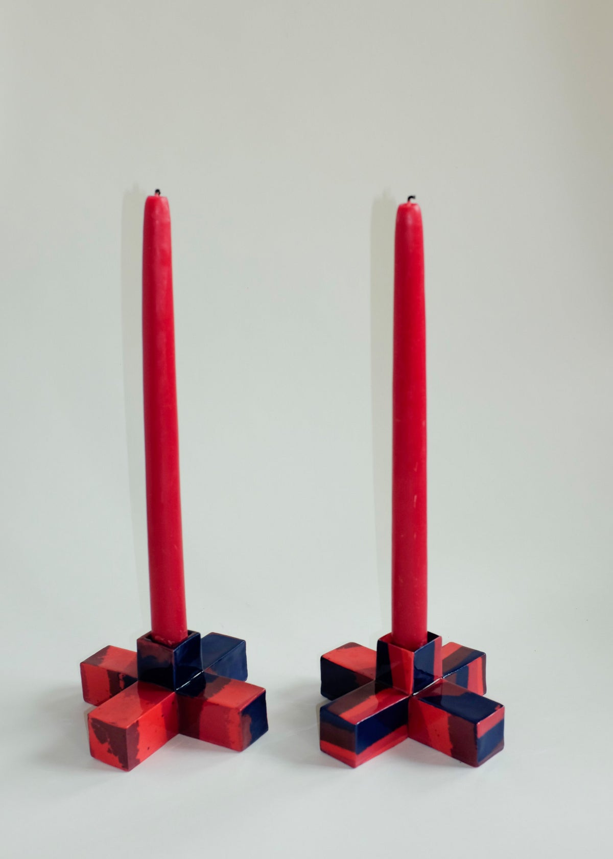 Two small squared candle holders in red and dark blue geometric patterns, each holding a tall red tapered candle against a plain light background.